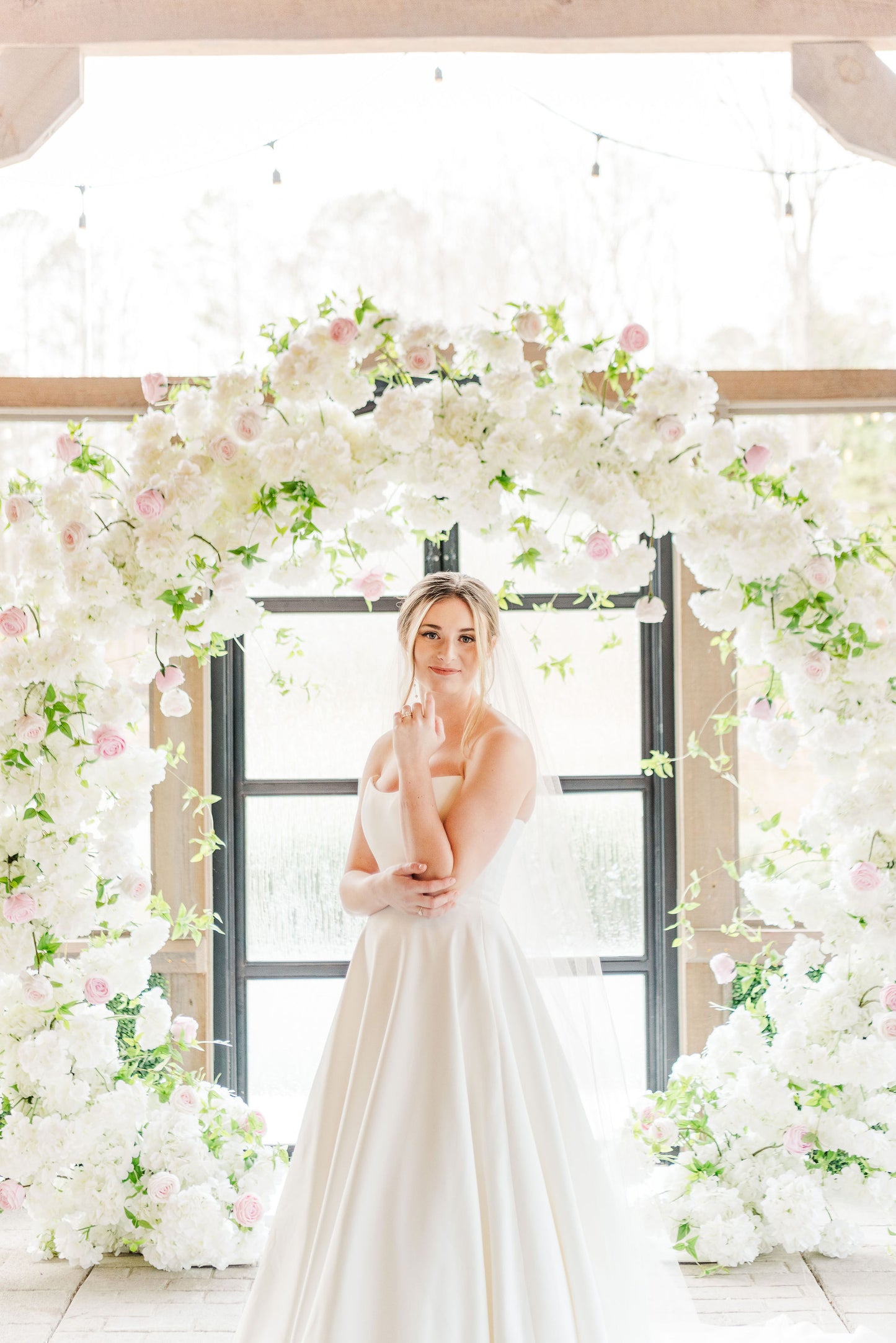 Bride beneath the Meadow Arch, framed by lush blush and ivory florals with greenery in a romantic outdoor setting.