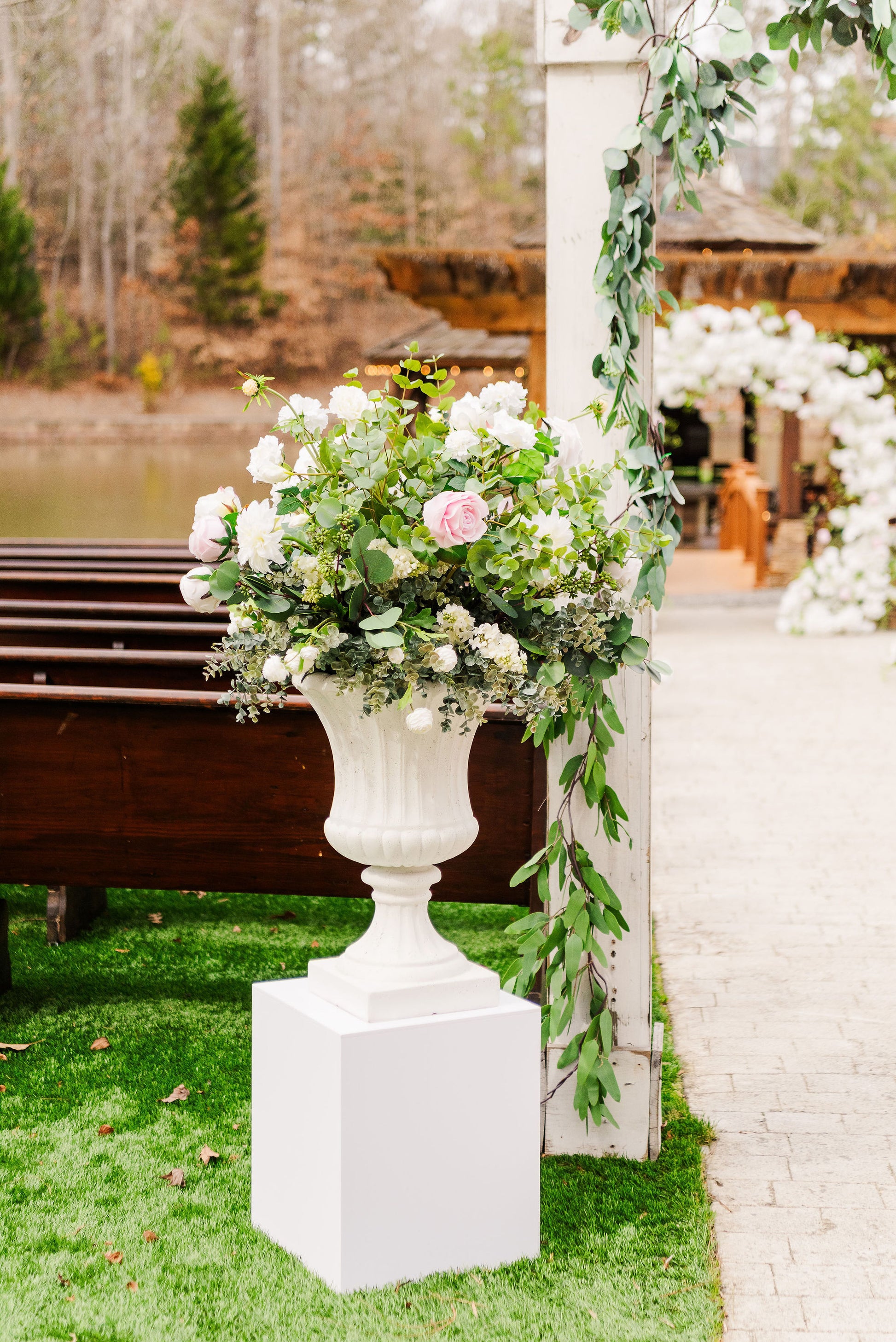 A white urn pedestal displayed in an outdoor setting with a floral arrangement on top.