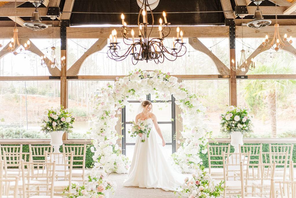 Wide view of the Meadow Arch as a focal point at the end of a floral-lined ceremony aisle styled with white chairs and soft colors.