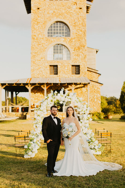 A bride and groom standing in front of a floral wedding arch with white and yellow flowers.