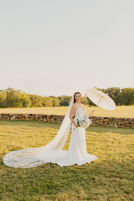 A woman in a white bridal gown holding a white parasol, standing in an outdoor setting.