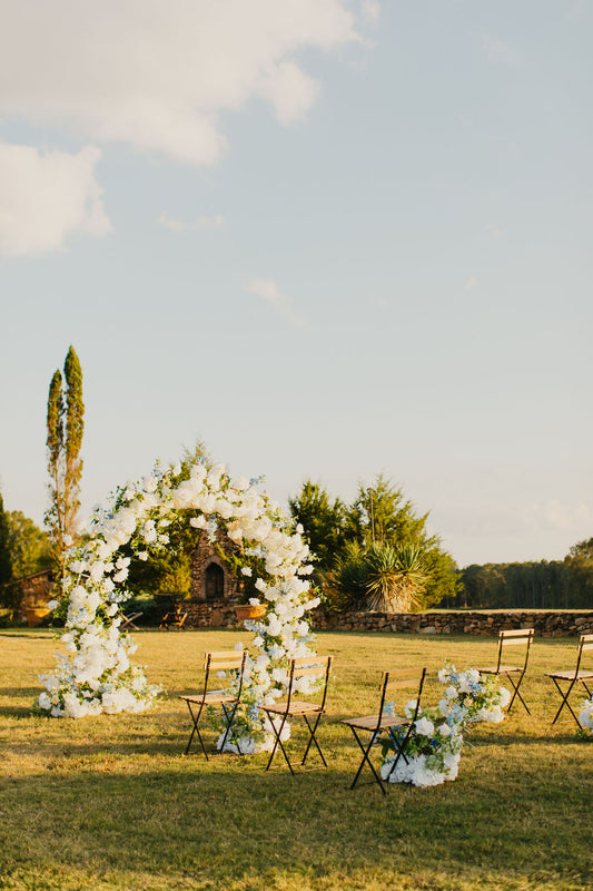 A wedding aisle with floral bouquets on either side, leading up to a wedding arch, with a backdrop of trees and clear sky.