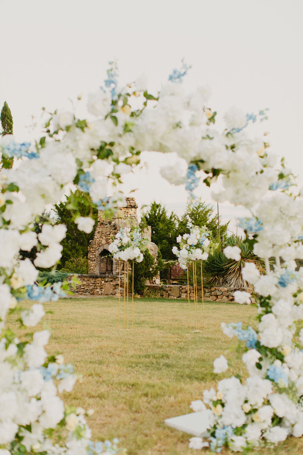 A floral arch with white and yellow roses, hydrangeas, delphinium, eucalyptus, and clematis set up outdoors for a wedding ceremony.