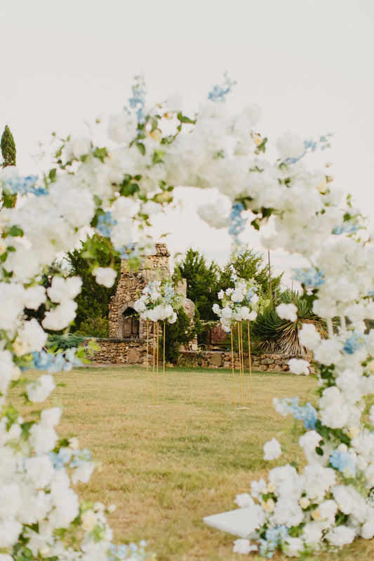 A floral arch with white and yellow roses, hydrangeas, delphinium, eucalyptus, and clematis set up outdoors for a wedding ceremony.