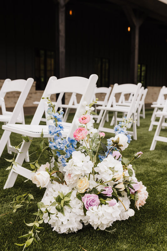 A vibrant wedding aisle bouquet with a variety of flowers including hydrangeas, roses, and other foliage, placed on the ground surrounded by white folding chairs.