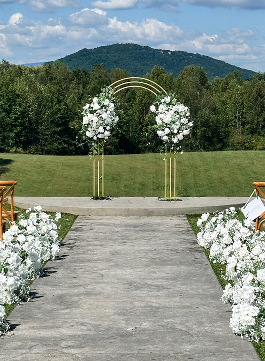 A golden wedding arch with lush floral sprays on each side, set against an outdoor backdrop with trees and mountains.