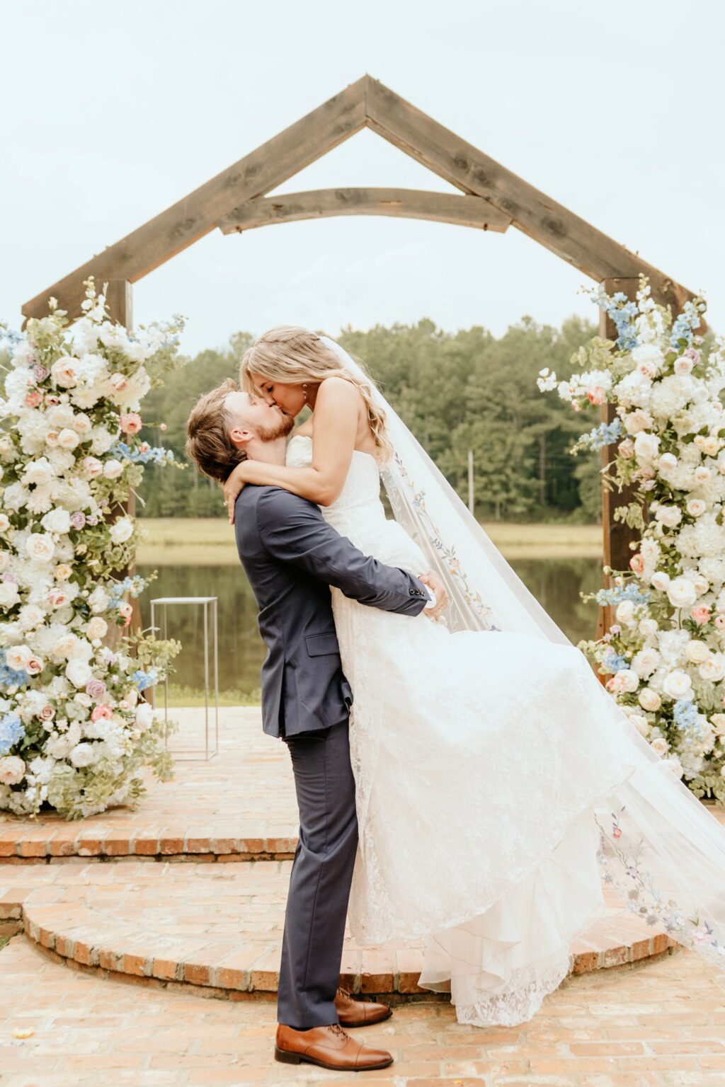Couple kissing beneath the Wildflower Wedding Pillars faux floral arch at a lakeside North Georgia wedding